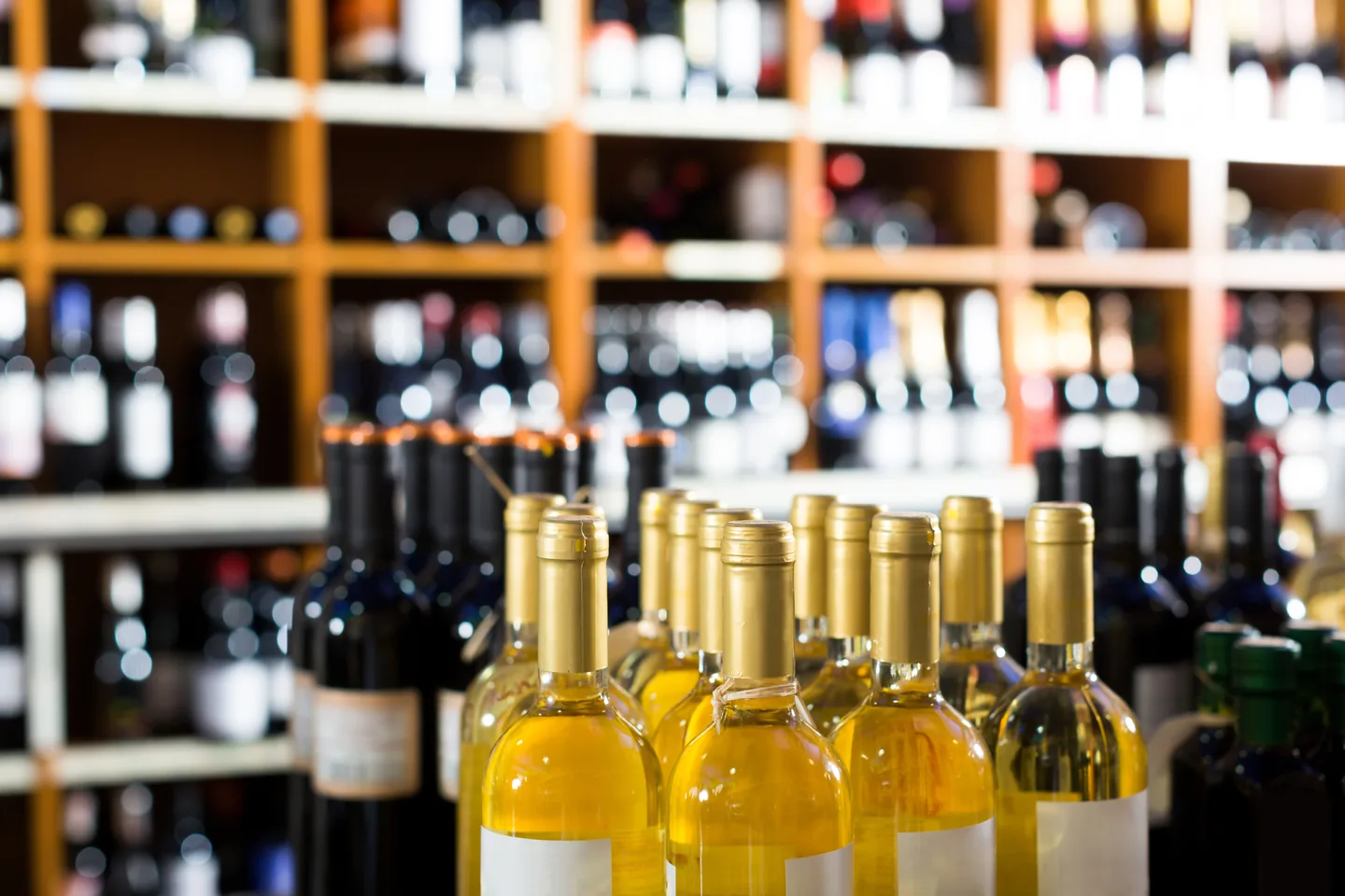 Gold-capped white wine bottles standing in front of wooden shelf cubbies stocked with wine, the background in soft focus