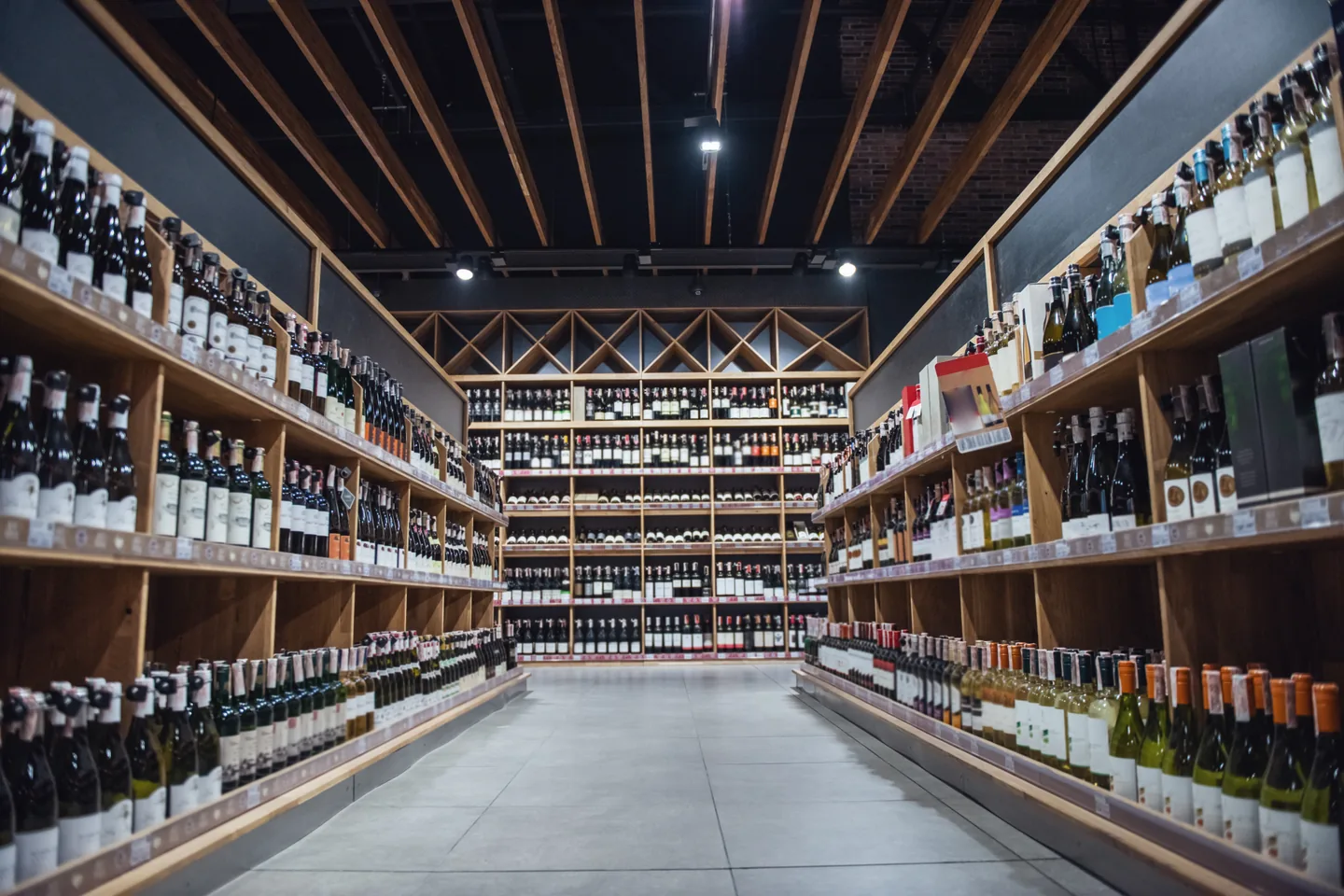 A retail aisle lined with stocked wooden wine shelving on both sides, lit by track lighting under exposed beams — liquor store insurance in Utah