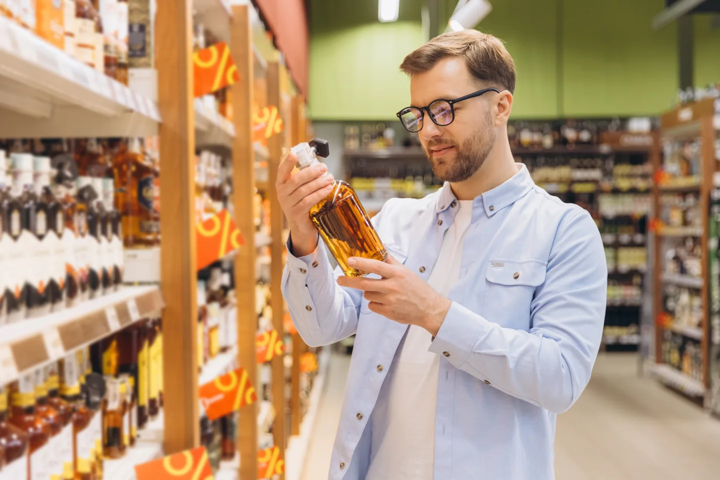 A customer in a retail aisle reading the label on a bottle of amber spirits, with discount tags along the shelf edges — liquor store insurance in Oklahoma