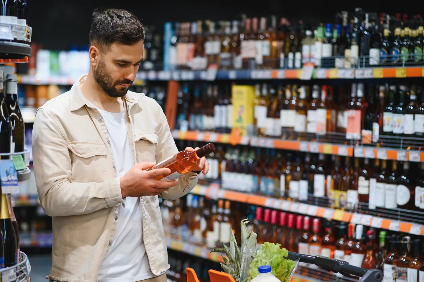 A shopper reading a bottle label beside a trolley of groceries, in front of a long aisle of wine and spirits — liquor store insurance in Nevada